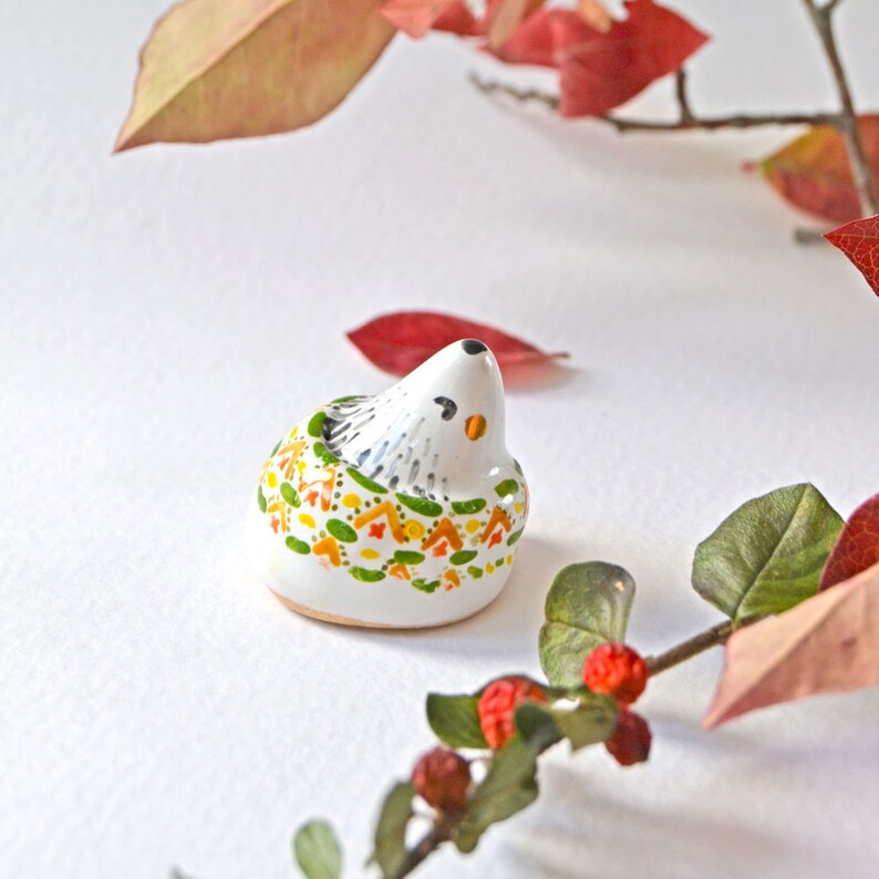 May include: A small, white ceramic hedgehog figurine with a black nose and eyes, and a decorative pattern of green, orange, and yellow leaves. The hedgehog is set against a white background with fall foliage.