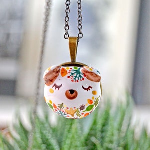 May include: A white ceramic pendant necklace with a cute animal face design. The pendant is decorated with colorful autumn leaves, berries, and branches. The necklace is on a silver chain.