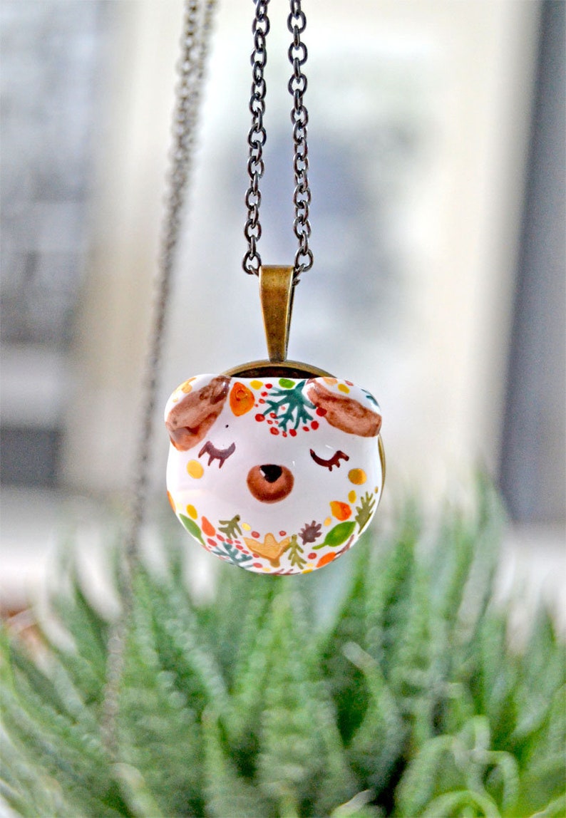May include: A white ceramic pendant necklace with a cute animal face design. The pendant is decorated with colorful autumn leaves, berries, and branches. The necklace is on a silver chain.