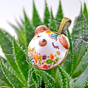 May include: A white ceramic pendant with a painted design of a bear's face and autumn leaves. The pendant is hanging from a gold chain and is resting on a green succulent plant.
