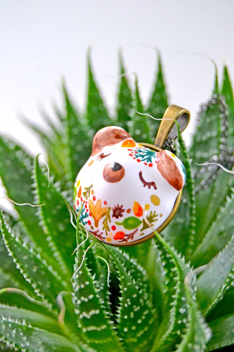 May include: A white ceramic pendant with a painted design of a bear's face and autumn leaves. The pendant is hanging from a gold chain and is resting on a green succulent plant.