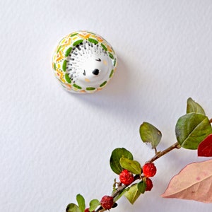 May include: A ceramic hedgehog figurine with a white body and a patterned design in green, yellow, and orange. The hedgehog has black eyes and a black nose. A sprig of leaves and red berries is in the bottom right corner.