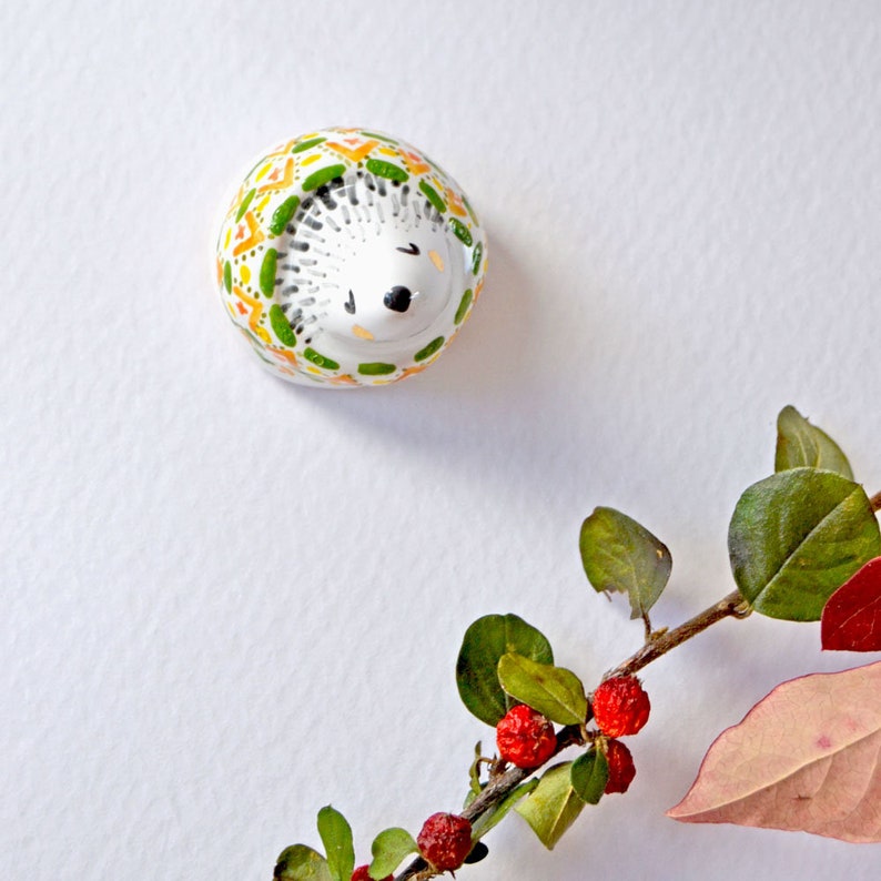 May include: A ceramic hedgehog figurine with a white body and a patterned design in green, yellow, and orange. The hedgehog has black eyes and a black nose. A sprig of leaves and red berries is in the bottom right corner.