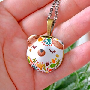 May include: A white ceramic pendant necklace with a hand-painted design of a bear's face with closed eyes and a floral pattern in shades of orange, yellow, green, and brown. The pendant is attached to a silver chain.