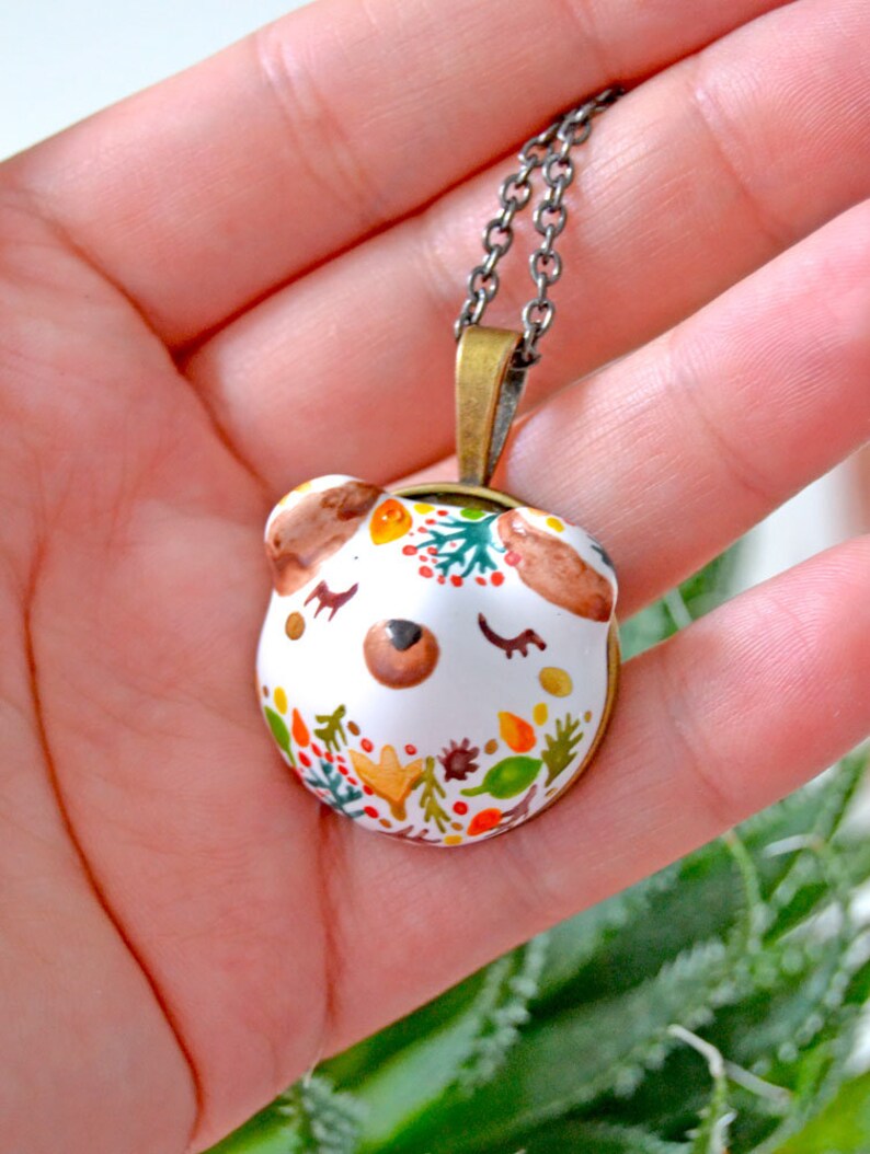 May include: A white ceramic pendant necklace with a hand-painted design of a bear's face with closed eyes and a floral pattern in shades of orange, yellow, green, and brown. The pendant is attached to a silver chain.