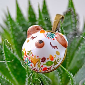 May include: White ceramic pendant with a painted design of a bear's face with closed eyes and a floral pattern in shades of red, orange, yellow, green, and brown. The pendant is attached to a bronze chain.
