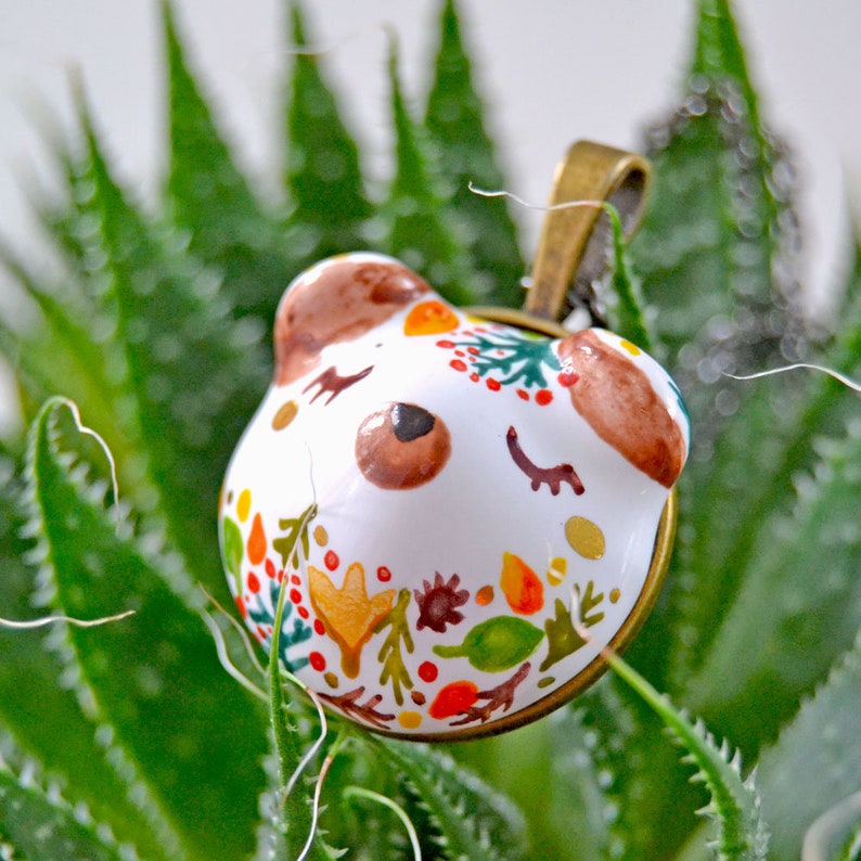 May include: White ceramic pendant with a painted design of a bear's face with closed eyes and a floral pattern in shades of red, orange, yellow, green, and brown. The pendant is attached to a bronze chain.