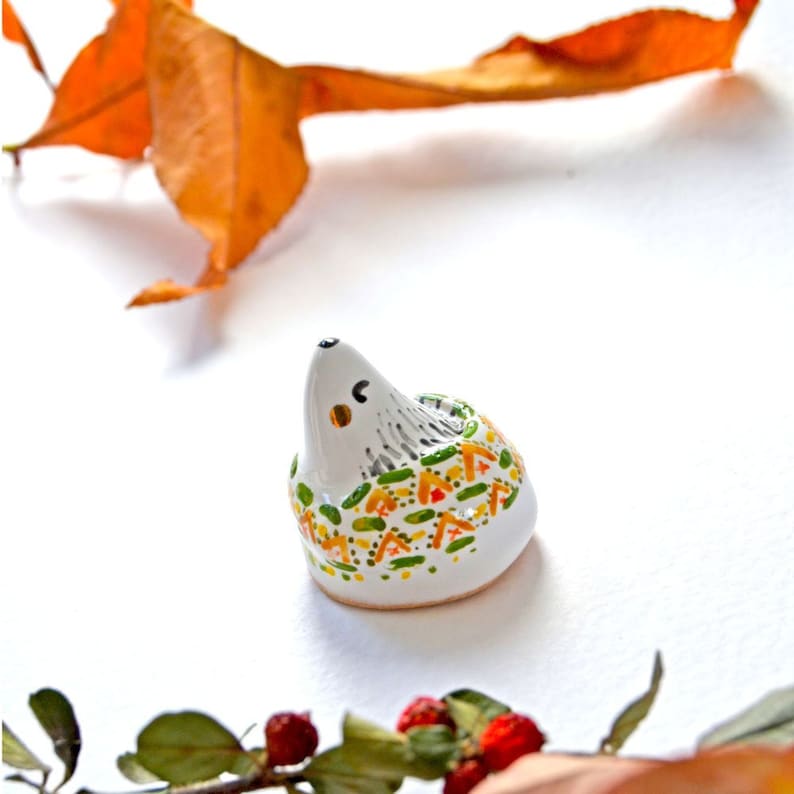 May include: A small, white ceramic hedgehog figurine with a painted design of green, orange, and yellow shapes. The hedgehog has a black eye and a small, orange ear. The figurine is on a white surface with a dried leaf and berries.