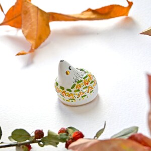 May include: A small, ceramic hedgehog figurine with a white body, gray spines, and colorful accents of green, yellow, and orange. The hedgehog has a black nose and small yellow eyes. The figurine is set against a white background with autumn leaves.