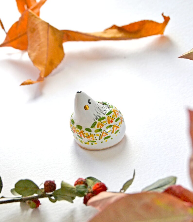 May include: A small, ceramic hedgehog figurine with a white body, gray spines, and colorful accents of green, yellow, and orange. The hedgehog has a black nose and small yellow eyes. The figurine is set against a white background with autumn leaves.