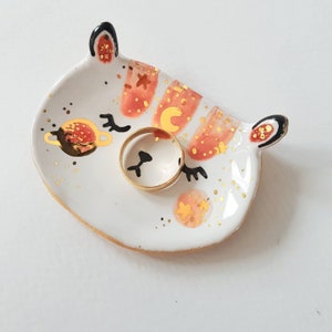 Op de afbeelding: Een witte keramische ringenschotel in de vorm van een kattenkop met gouden en oranje details. De schotel heeft een gouden ring erin.