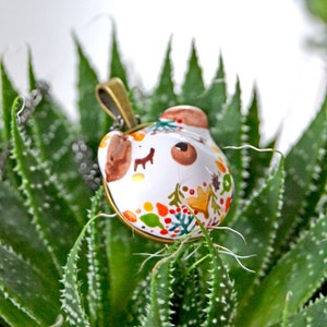 May include: A white ceramic pendant with a painted design of a bear face and autumn leaves in shades of red, orange, yellow, and green. The pendant is attached to a gold chain.