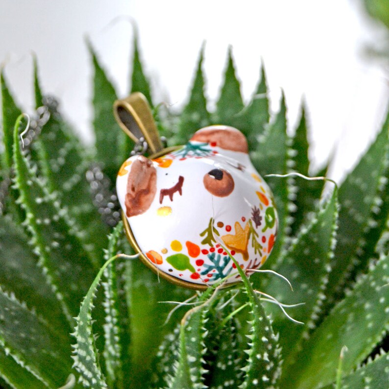 May include: A white ceramic pendant with a painted design of a bear face and autumn leaves in shades of red, orange, yellow, and green. The pendant is attached to a gold chain.