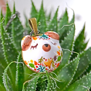 May include: A white ceramic pendant necklace with a hand-painted design of a bear's face with closed eyes. The bear's face is surrounded by a colorful floral pattern with red, orange, yellow, green, and brown accents. The pendant is attached to a silver chain.
