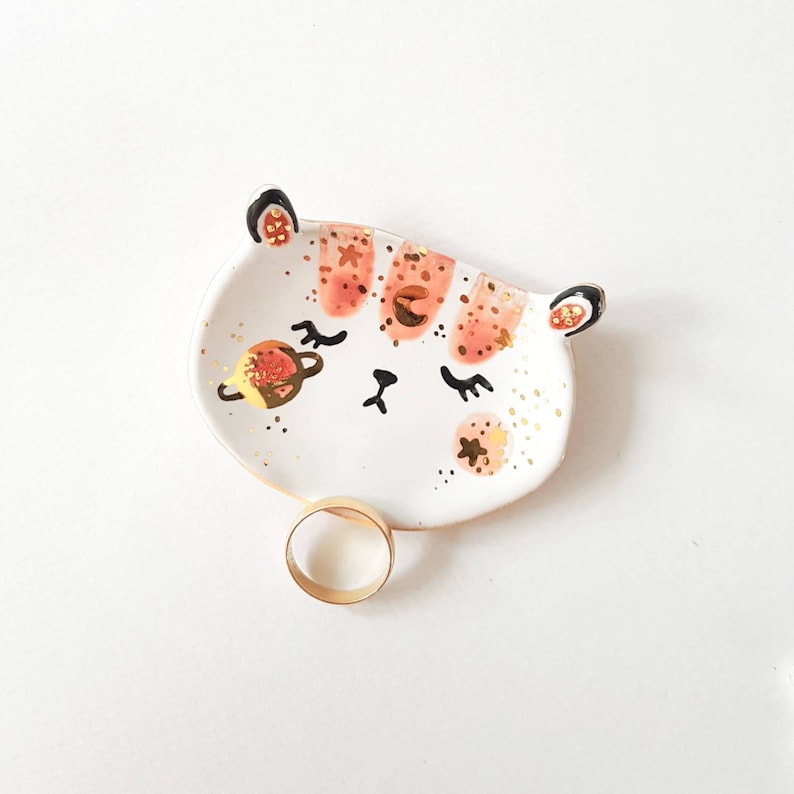 Op de afbeelding: Witte keramische ringenschotel met een handgeschilderd ontwerp van een kattenkop met roze strepen, gouden glitter en een gouden planeet.