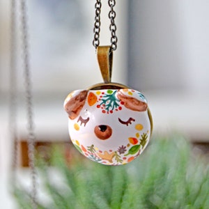 May include: A white ceramic pendant necklace with a whimsical animal face design. The pendant features a brown nose, closed eyes, and colorful floral and leaf accents. The necklace is on a bronze chain.