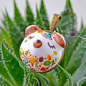 May include: A white ceramic pendant with a brown bear face and colorful floral designs. The pendant is attached to a brown metal chain.