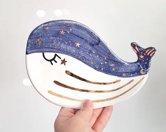 May include: A white ceramic whale dish with a blue and gold design. The whale has a closed eye with a gold star above it. The whale's body is decorated with gold stars and a crescent moon.