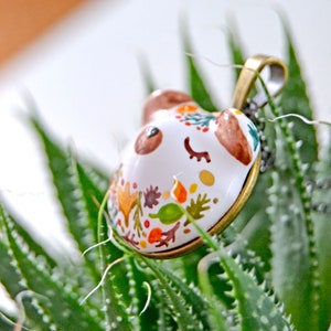 May include: A white ceramic pendant with a painted design of leaves and flowers in shades of orange, yellow, green, and brown. The pendant is shaped like a bear's head and has a gold-toned metal bail.