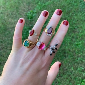 May include: A hand displaying five rings with colorful gemstones. The rings feature a variety of designs, including a ruby, emerald, and a pink rectangular stone. The rings are set in gold and silver bands.