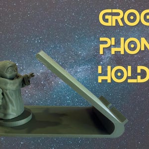 Puede incluir: Un soporte para teléfono Grogu de color gris verdoso. El soporte presenta una figura de Grogu con los brazos extendidos y un soporte inclinado para un teléfono. El fondo es un cielo estrellado. El texto "GROGU PHONE HOLDER" es amarillo.