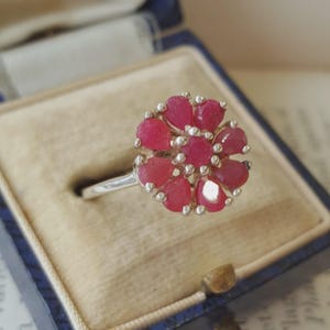 Può includere: Un anello in argento con un gruppo di pietre preziose a forma di pera di colore rosso rosato intenso, disposte in un motivo floreale attorno a una pietra centrale rotonda. La montatura è in argento metallico e le pietre sono sfaccettate.