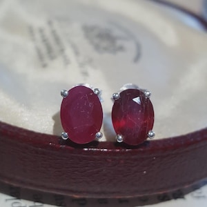 Può includere: Un paio di orecchini a lobo con rubini rossi di forma ovale montati su griffe in metallo color argento. Ogni gemma presenta una superficie sfaccettata che riflette la luce, mettendo in risalto una profonda tonalità cremisi.