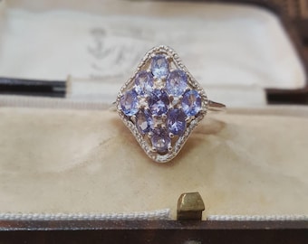 May include: A silver ring featuring a cluster of oval-cut, light purple gemstones set in a diamond-shaped design. The ring is displayed inside a vintage jewelry box with a beige interior.