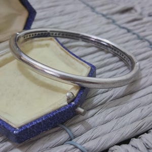 Peut inclure: Un bracelet jonc argenté avec une rangée de petites pierres transparentes serties sur le bord supérieur. Le bracelet est présenté dans un écrin bleu avec un intérieur crème. La boîte est ouverte, révélant le bracelet.