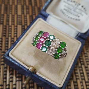 May include: A silver ring set with a cluster of gemstones. The ring has pink, green, and clear stones. It is presented in a blue jewellery box with a cream-coloured interior.