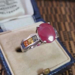 Peut inclure: Une bague argentée ornée d'une grande pierre précieuse rouge foncé de taille ovale en son centre. L'anneau est agrémenté d'une rangée de pierres plus petites de taille carrée, déclinées en bleu, orange, jaune et vert, serties dans le métal.