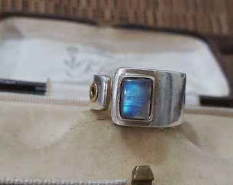 May include: A silver ring with a rectangular, iridescent blue gemstone. The ring has a unique design with a smaller, adjacent element featuring a gold accent. The ring is displayed in a vintage jewelry box.