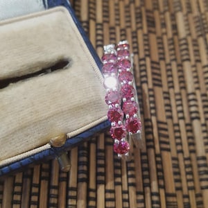 Peut inclure: Une paire de boucles d'oreilles en argent avec une rangée de pierres précieuses roses vives. Les boucles d'oreilles sont présentées dans un écrin bleu avec un intérieur beige. Les pierres sont rondes et espacées.