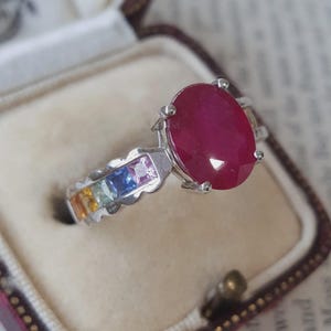 May include: A silver-toned ring featuring a large, oval-cut, deep red gemstone as the centrepiece. The band is accented with a series of smaller, square-cut gemstones in a gradient of colours, including pink, blue, yellow, and orange hues.