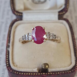 Può includere: Un anello in oro con un rubino centrale rosso intenso dal taglio ovale, fissato da quattro griffe. Il gambo è decorato su entrambi i lati da una disposizione a gradini di tre piccoli diamanti trasparenti dal taglio baguette, creando un design simmetrico.