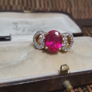May include: A gold ring featuring a large, oval-cut, bright pink gemstone. The ring is accented with two gold sections, each set with small, clear gemstones. The ring is presented inside a vintage jewellery box.