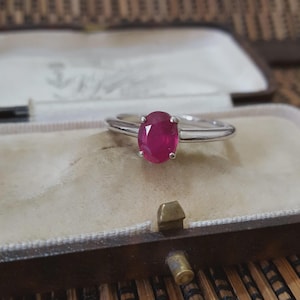 May include: A silver ring featuring a large, oval-cut ruby gemstone. The ruby is a deep red hue, set in a classic four-prong setting. The ring is presented in a vintage jewellery box with a beige interior.