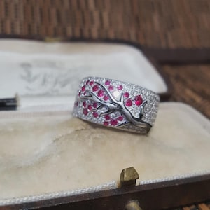 May include: A wide silver ring with a tree branch design and red gemstone blossoms. The band is encrusted with small, clear gemstones. The ring is displayed in a vintage jewellery box.