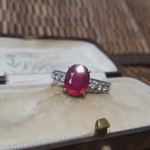 Op de afbeelding: Een zilveren ring met een grote ovale robijn in het midden en kleinere heldere stenen langs de band. De ring wordt gepresenteerd in een vintage sieradendoos met een crèmekleurig interieur.