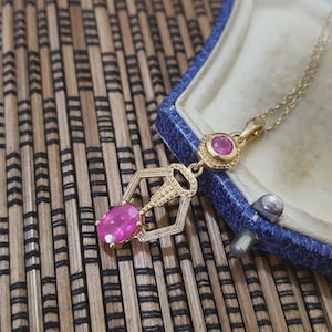May include: A gold pendant necklace featuring a large, oval, pink gemstone and two smaller pink gemstones. The necklace is presented in a blue jewellery box against a textured, brown background.