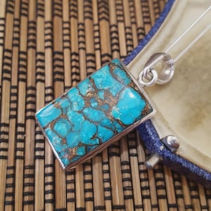 May include: A rectangular pendant featuring a turquoise and brown stone set in a silver frame, hanging from a fine silver chain. The stone displays a distinctive, natural pattern. The pendant is presented in a blue jewellery box.