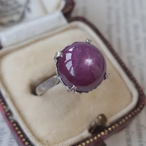 Può includere: Un anello in argento con una grande pietra preziosa rubino stellato di colore magenta intenso, tagliata a cabochon rotondo. La pietra è fissata da quattro griffe metalliche che si estendono dalla fascia, caratterizzata da un aspetto organico.