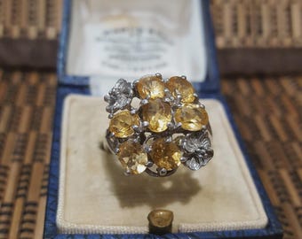 May include: A silver ring featuring a cluster of oval-cut, golden-yellow gemstones. The ring has a floral design on the sides and is set in a vintage blue jewelry box with a cream-colored interior.