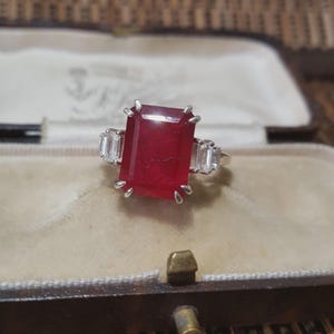 May include: A silver ring with a large, rectangular, deep red gemstone, and two smaller, rectangular, clear gemstones. The ring is set in a vintage style, displayed in a velvet-lined jewellery box.