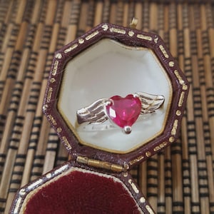 Può includere: Un anello d'argento con una pietra a forma di cuore color rubino e dettagli alati. L'anello è presentato in un portagioie ottagonale marrone aperto, con interno color crema, su uno sfondo di legno naturale.
