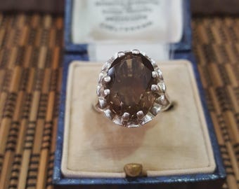 May include: A vintage silver ring with a large, oval-cut smoky quartz gemstone. The ring is set in a decorative, openwork setting and is displayed inside a blue jewelry box with a cream-colored interior.