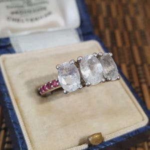Peut inclure: Une bague argentée ornée de trois pierres blanches transparentes de taille ovale alignées. L'anneau est agrémenté d'une série de petites pierres rondes rose vif sur le côté, créant un contraste avec les pierres centrales transparentes.