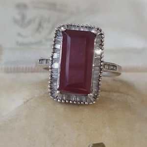 Op de afbeelding: Een zilverkleurige ring met een grote, rechthoekige, donkerrode steen in het midden. De steen is omringd door een halo van kleine, heldere baguette-geslepen stenen in een metalen rand met kraalmotief. De ringband bevat ook kleine steentjes.