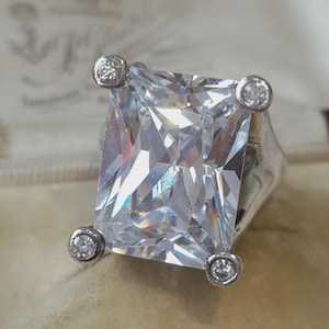 May include: A silver-toned ring featuring a large, rectangular, faceted clear stone as the centrepiece. The stone is secured by four prongs, each topped with a small, round, clear accent stone, creating a distinct four-cornered setting design.