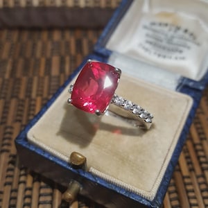 Könnte beinhalten: Ein silberner Ring mit einem großen, rechteckigen, roten Edelstein. Das Band ist mit kleinen, klaren Steinen besetzt. Der Ring wird in einer blauen Vintage-Schmuckschatulle mit beigem Innenraum präsentiert, was ein klassisches und elegantes Design zeigt.